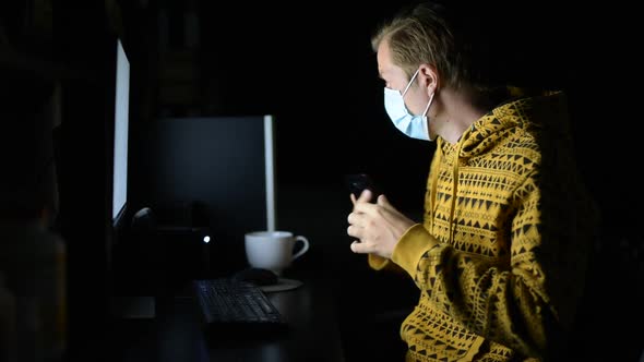 Young Man with Mask Using Phone While Working From Home Late at Night in the Dark