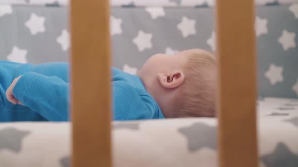 Newborn Boy in Azure Bodysuit Moves Hands Resting in Crib