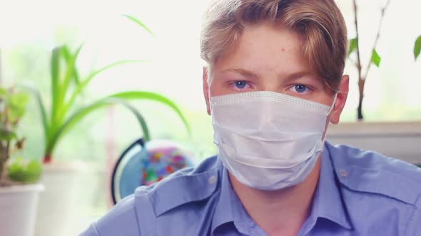  Teenager of European Appearance in a Blue Shirt Sits at Home, Puts on a Medical Protective Mask