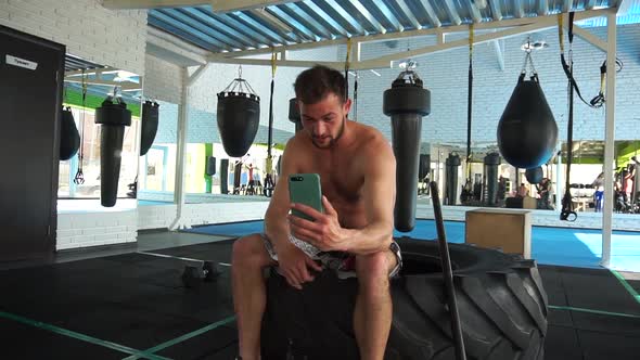 Young Man Sitting in the Gym, Rest After Workout, Using Smartphone, Speaking with Subscribers