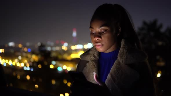 A Young Black Woman Works on a Smartphone in an Urban Area at Night  City Lights in the Background