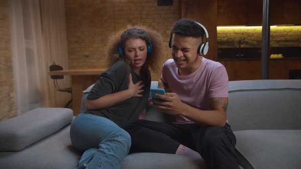 20s Mixed Race Couple Wearing Headphones Singing Songs Looking at Mobile Phone Sitting on Couch