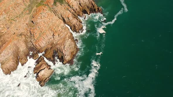 Teal ocean waves breaking onto shoreline with jagged rocky cliff. View from a drone.