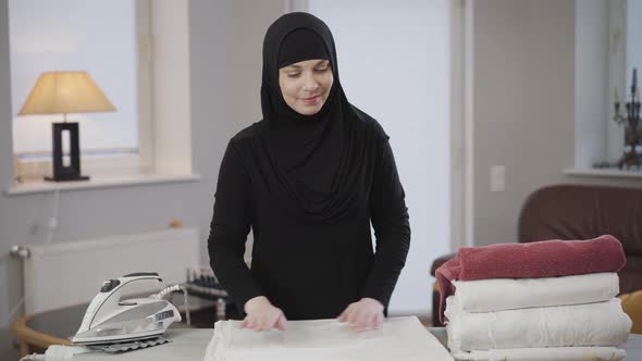 Middle Shot Portrait of Smiling Muslim Woman Standing in Front of Ironing Board with Laundry and