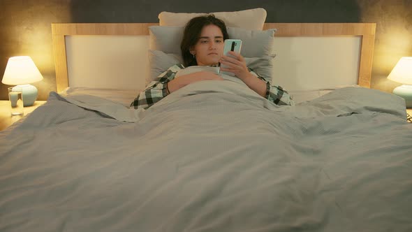 Brunette Girl Lying in Bed in the Evening and Reading Messages on Her Cell Phone Front View