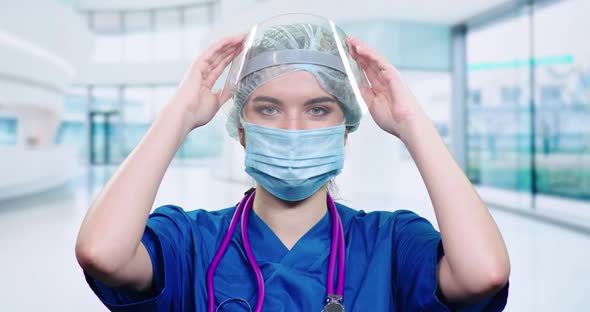 Portrait of Young Doctor Female in Protective Gear and Medical Mask, Woman Puts on a Plastic Visor