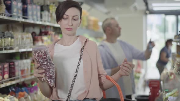 Focused Young Woman Putting Packaging of Raw Beans Into Shopping Basket, Blurred Man Choosing