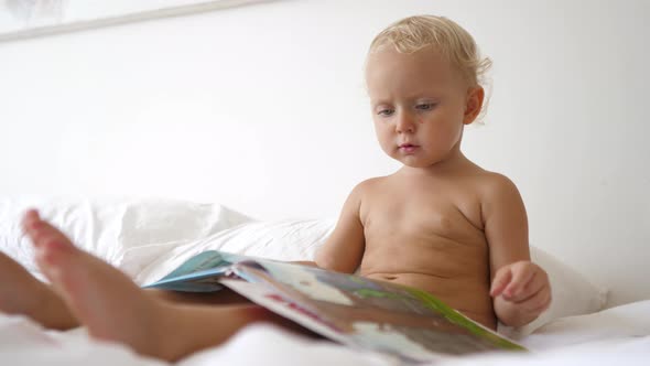 Cute Blonde Toddler Reading a Colorful Book to Himself in Bed