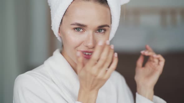 Portrait of Woman in Bathrobe, with Towel on Head Applies Cream on Her Face
