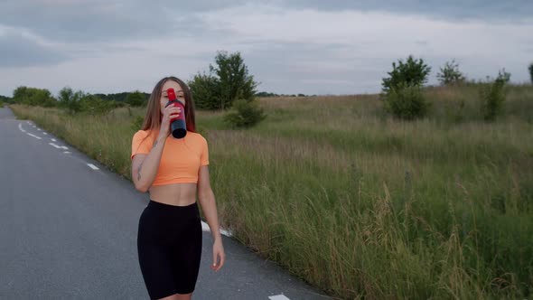 Young Athletic Girl Walking on a Road in Morning and Drinking From Sport Bottle