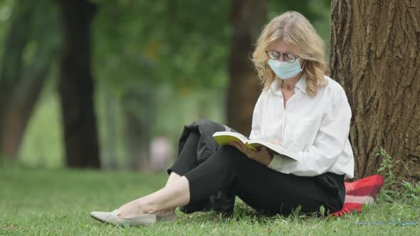 Engrossed Middle Aged Blond Woman in Eyeglasses and Coronavirus Face Mask Reading Book Sitting on