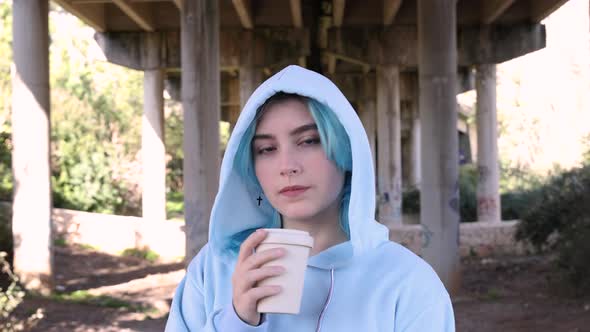 Blue haired teenage girl in oversize hoodie drinking coffee to go against bridge pillars