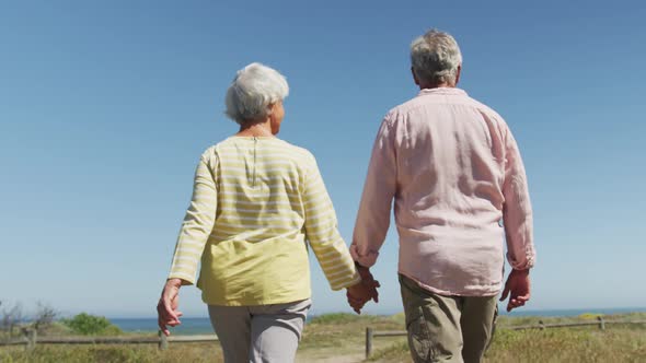 Senior caucasian couple holding hands and walking on path leading to the beach