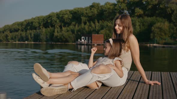 Woman with Daughter Sit on a Pier Looking at Lake Smiling at the Sunset