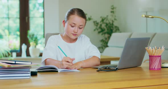 Cute Teen Girl Sits at the Table with Laptop Does Her Homework Writing with Pencil in Notebook Books alt