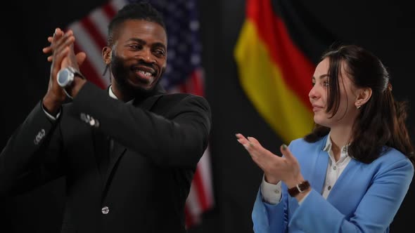 Clapping Caucasian Woman Inviting African American Man to the Stage in Camera Flashes at Press