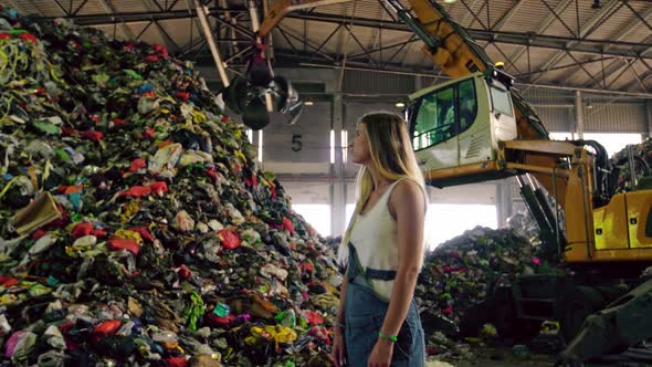 Young Woman in Jeans Standing Against Operating Excavator Against Huge Pile of Trash