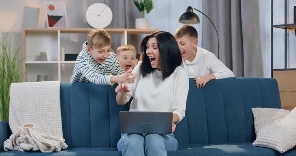 Woman Sitting on the Sofa and Uses Laptop when Her Playful Carefree three Sons Jumping Out