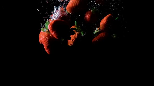 Strawberries dropped into water on black background, side lit slow motion