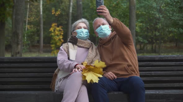Elderly Couple in Medical Masks Sits on a Bench in a Forest Park and Take a Selfie Video