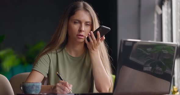 Woman Sitting in Cafe Holding Pen and Making Notes in Diarylaughingtalking on Speaker Using Phone