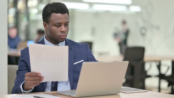 African Businessman Reading Documents While Working on Laptop