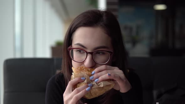 A Young Woman Is Eating a Burger. A Girl Sits in a Cafe By the Panoramic Window and Eats Fast Food.