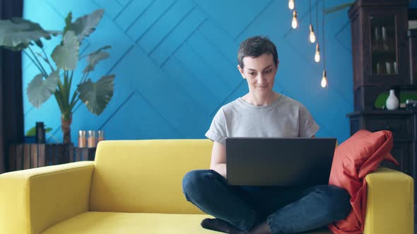 Woman with short hair is sitting on a yellow sofa with legs folded and typing on a laptop. freelance