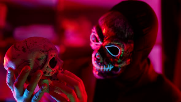 a Man Out of Focus and Wearing a Scary Glowing Mask Holds a Skull in Front of Him and Carefully