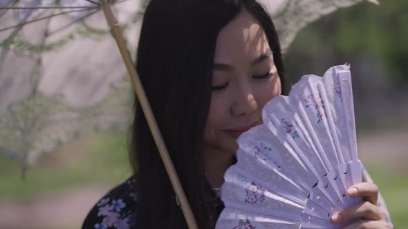 Closeup of Charming Shy Asian Young Woman with White Sun Umbrella Shaking Fan Smiling