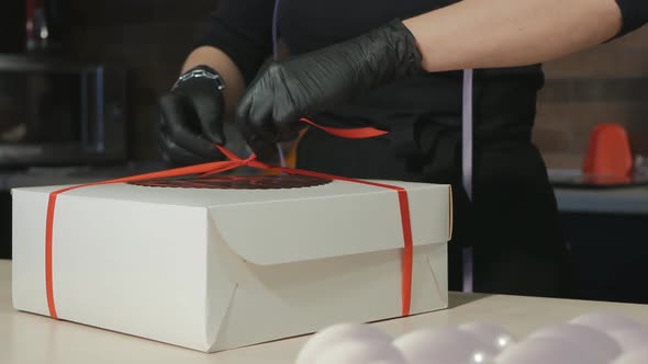 Woman Pastry Chef in a Black Apron and Black Gloves Is Tying a Ribbon on the Cake Package