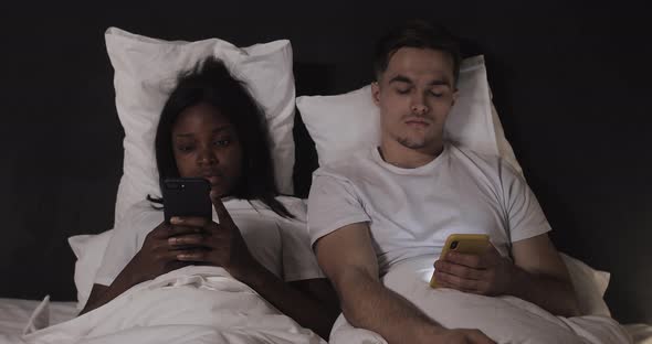 Multi-ethnic Couple Using Smartphones Lying in the Bed at Night. Man Peeks on His Woman s Phone, She