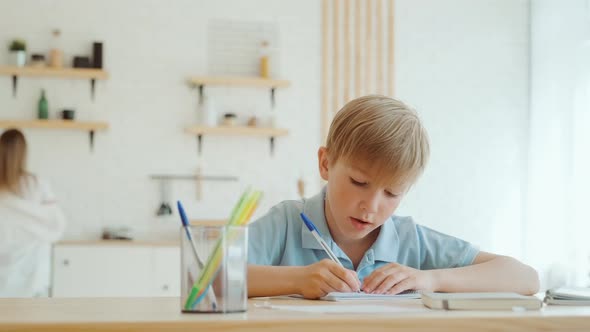 Little Boy is Doing Homework at Home Sitting at the Table Writing in Notebook