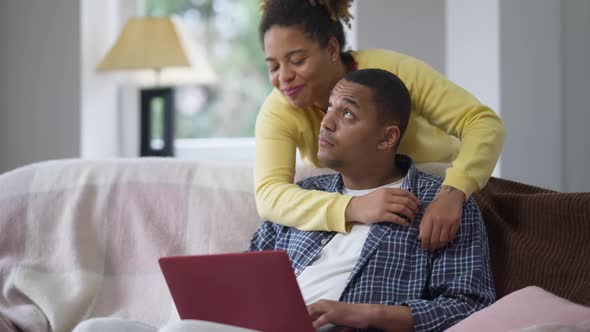 Busy Absorbed African American Man Sitting on Couch Messaging Online As Loving Woman Hugging Husband