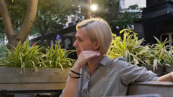 Cute Caucasoid Blonde Adult Scratching Her Nose with Her Finger While Sitting in a Street Restaurant