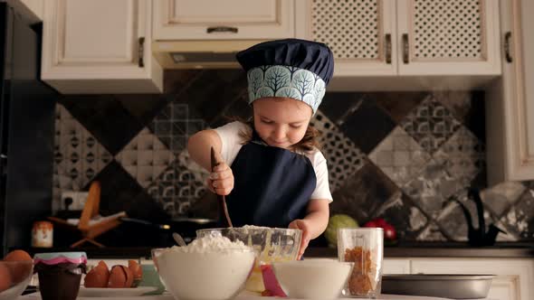 Little Girl in a Cook's Cap in the Kitchen, She Ineptly Mixes the Dough in Plate