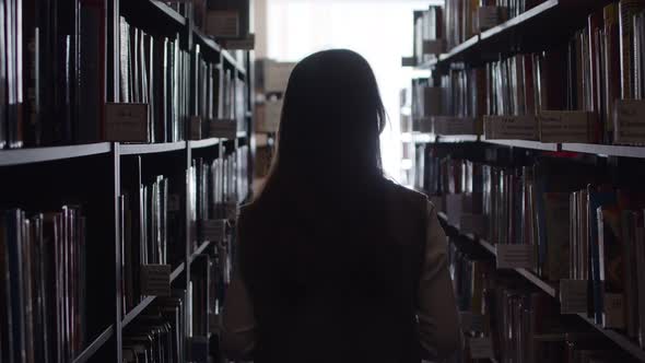 Woman in a sweater with long hair in the library, looking for a book among the shelves, Book Archive
