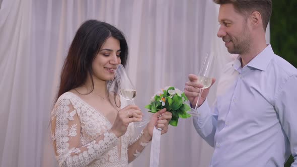 Side View of Happy Newlyweds Clinking Glasses Drinking Champagne at Wedding Altar Outdoors