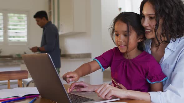 Happy hispanic mother and daughter sitting at table looking at laptop