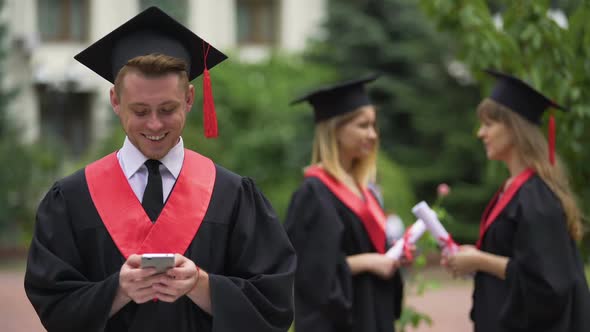 Young Smiling Man in Graduation Cap and Mantle Texting on Smartphone, Mobile App