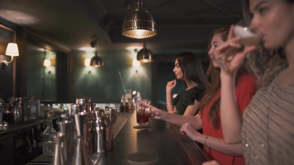 Three Beautiful Young Girls in Evening Dresses Sitting at the Bar Counter and Drinks Cocktail