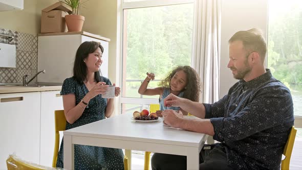 Beautiful Family with Kid Sitting at the Table Drinking Coffee at New Home Around Cardboard Boxes