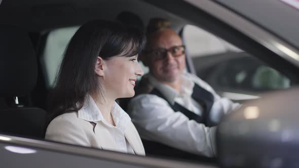 Side View Portrait of Happy Adult Wife Talking with Husband Sitting in New Car in Dealership Turning