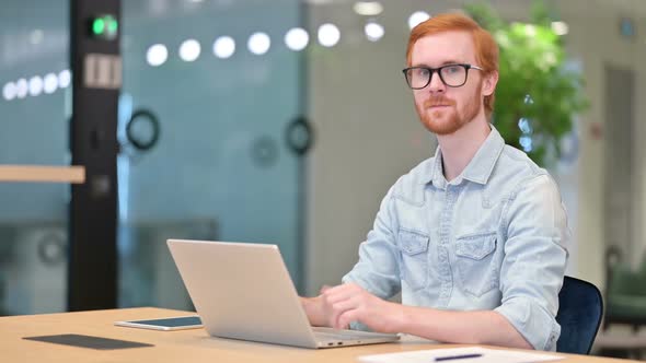 Thumbs Up By Young Redhead Man Working on Laptop in Office