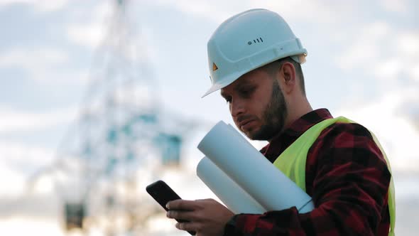 Worker Male Engineer Using Mobile Phone for Checking Data While Standing Against High Voltage Power