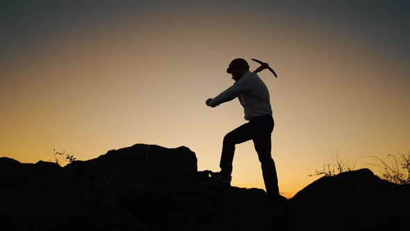 Silhouette Man in Helmet with Pickaxe in His Hands Stands on Rock and Hits Stone Side View