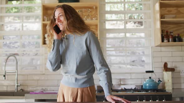 Happy caucasian woman standing in sunny cottage kitchen talking on smartphone and smiling