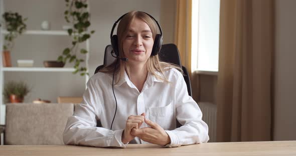 Portrait of Young Woman Sitting at Table at Home or in Office, Wears Headphones, Speaks with Clients