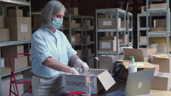 Mature Female Warehouse Worker Wearing Mask Packing Donations Box