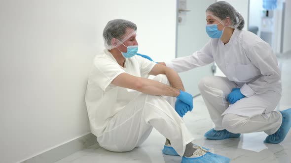 Depressed Caucasian Man Sitting on Floor in Hospital Corridor Leaning on Wall As Colleague Touching